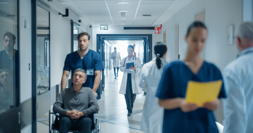 Busy Hospital Hallway Filled with Medical Experts in White Coats and Blue Uniforms. Middle Aged Female Doctor Reviewing a Patient's History on a Tablet Computer as She Heads to the Examination Room
