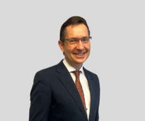 A head shot of a smiling brunette man wearing glasses and a suit with a red patterned tie