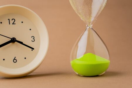 a clock and an hourglass with green sand stood next to each other on a beige background.