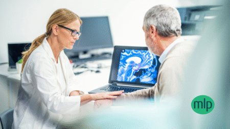 A doctor in a white lab coat and glasses, sat across from an older patient in a beige jumper. In front of them is a brain scan with a section highlighted with a small purple circle.