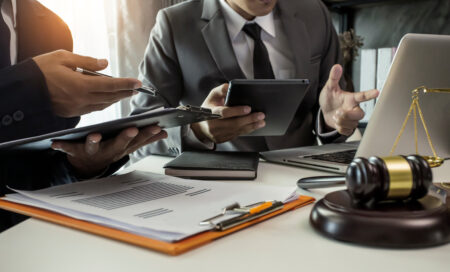 Two male lawyers sat at a table. Both are wearing suits and holding clipboards with a gavel on the table in front of them.