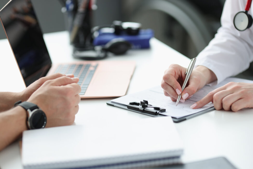 A GP sat across a desk from a patient. Only their hands and arms are visible in the shot with the doctor writing notes on a clipboard.